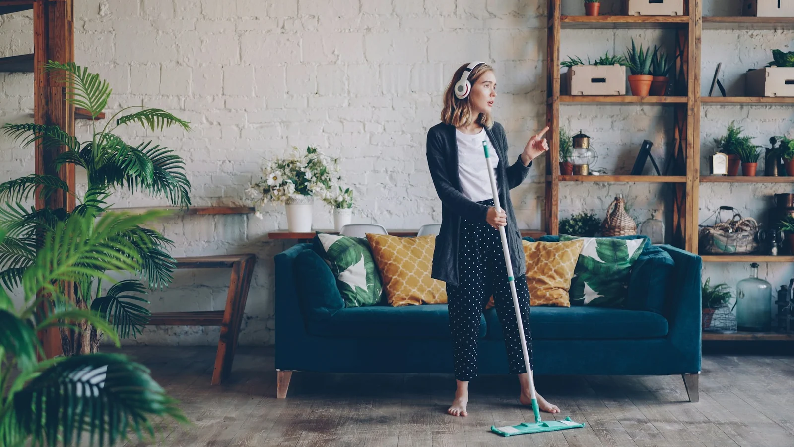 Woman in headphones mopping a wood floor beside a teal sofa and plant-filled shelves