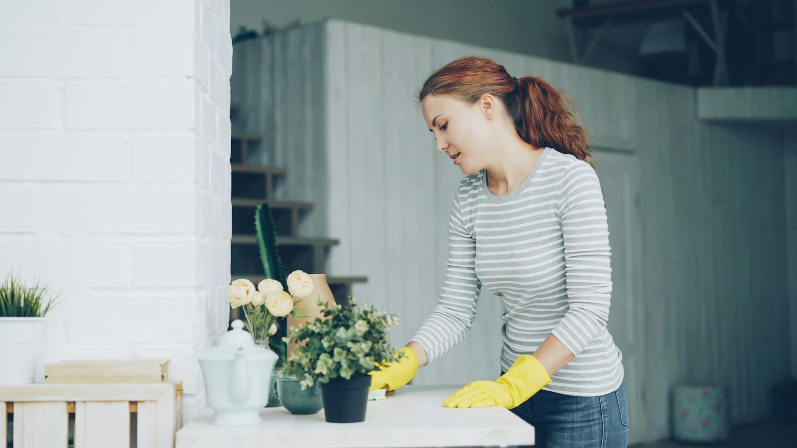 Woman in yellow rubber gloves wiping a white table next to potted plants