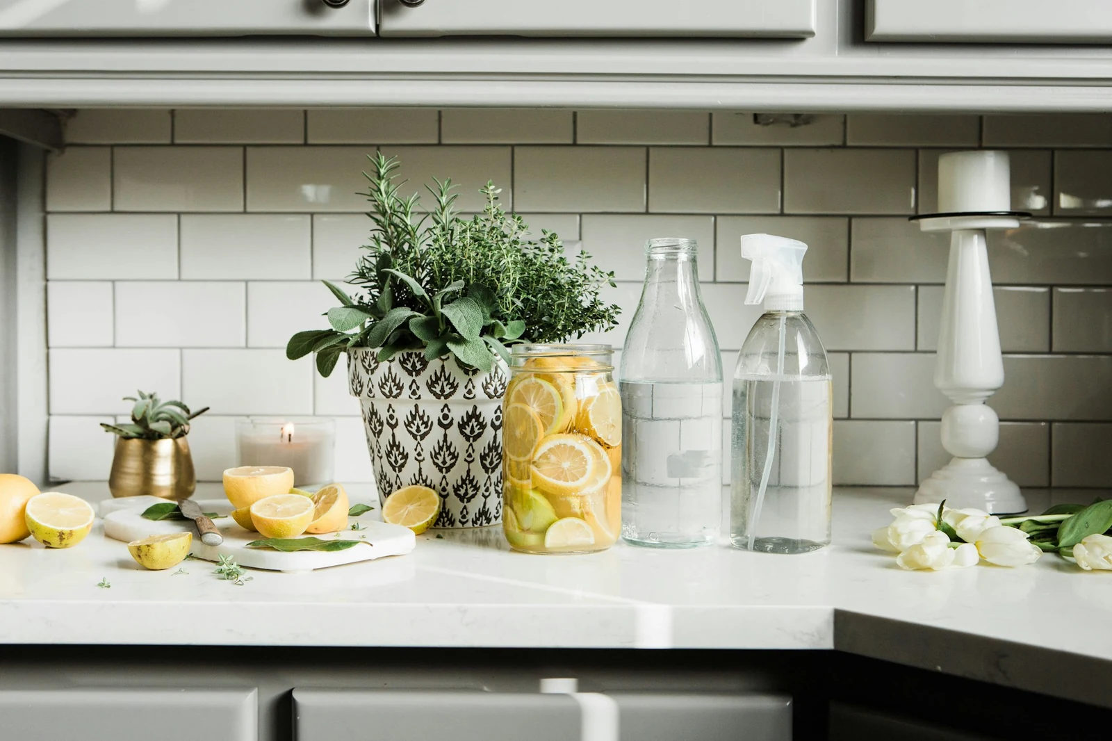 Glass spray bottle, jar of lemon slices and fresh herbs on a white kitchen counter